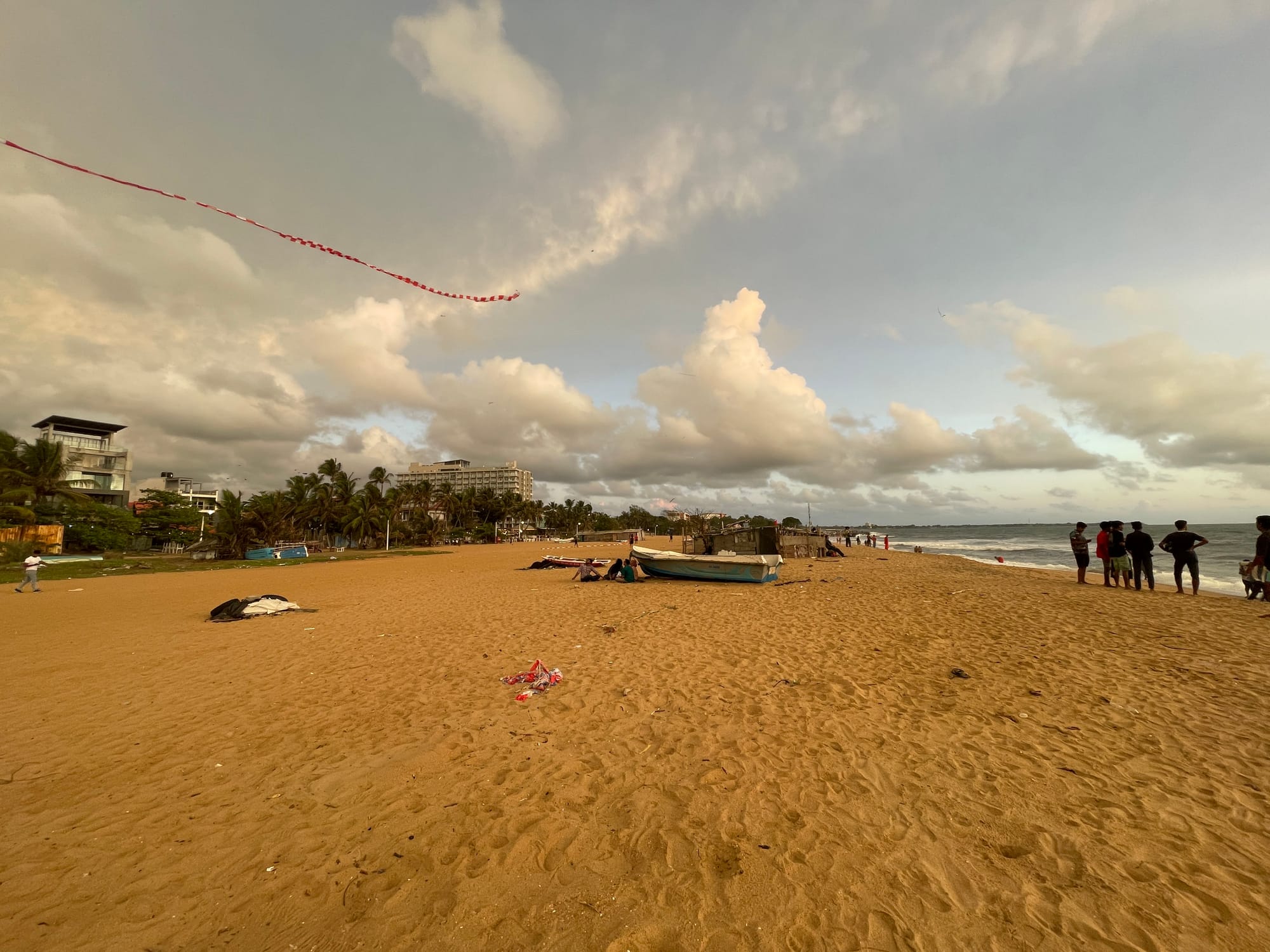 Playa de Negombo, Sri Lanka, con arena dorada, botes de pesca, personas cerca del mar y una larga cometa roja bajo un cielo nublado, por el fotógrafo de viajes Vincent Croos