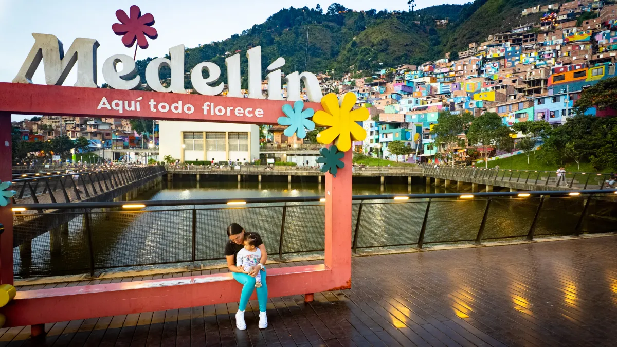 Letrero de Medellín (Manrique, Comuna 3) frente a casas coloridas en la ladera, con una mujer y una niña sentadas junto al paseo peatonal.