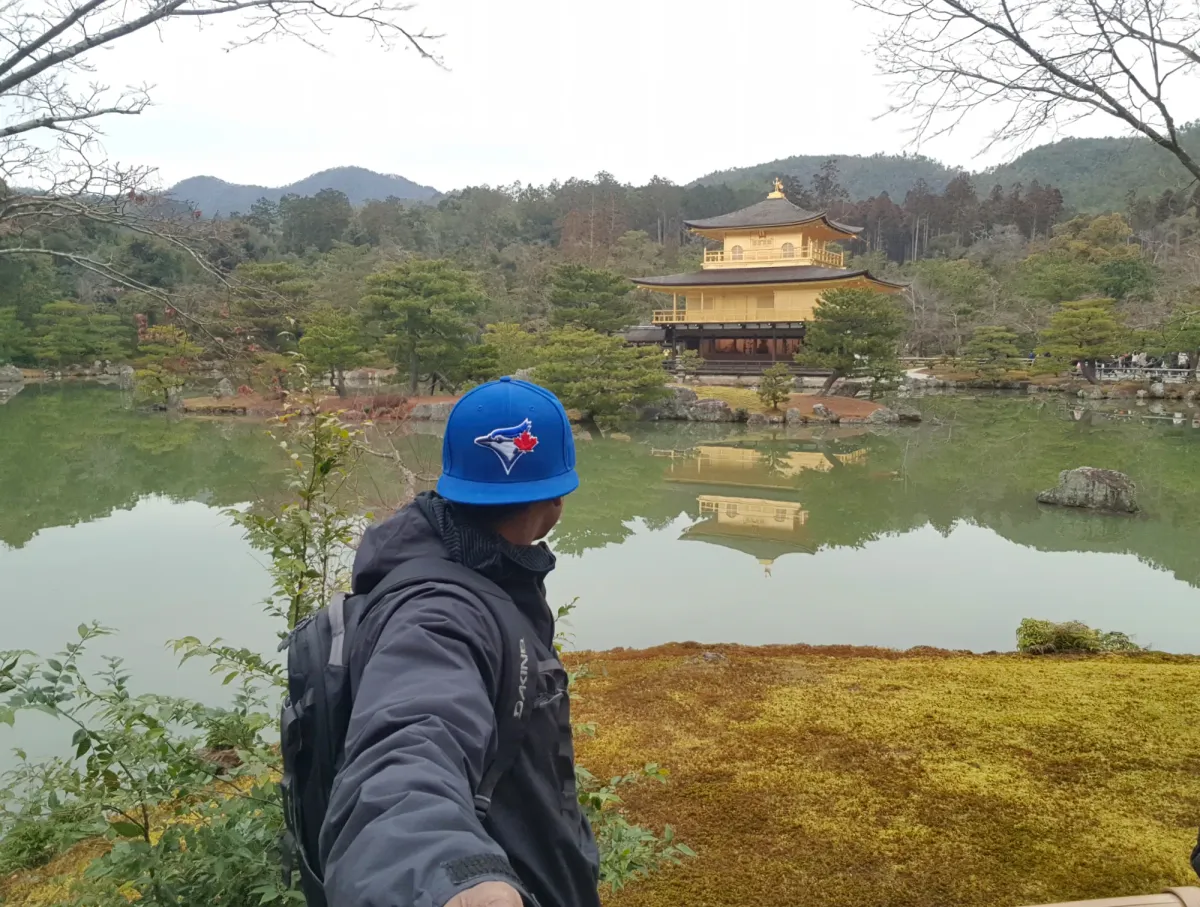 Hombre con gorra azul mirando el Pabellón Dorado y su reflejo en un estanque en Kioto, Japón.